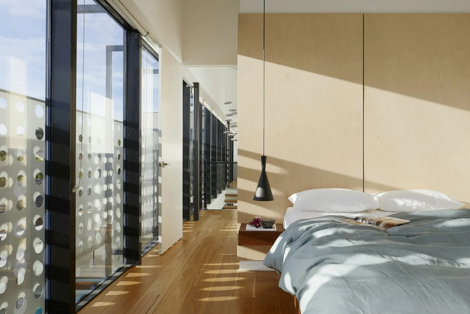 Daylit bedroom with warm timber flooring, a simple bed, and a glass-lined hallway