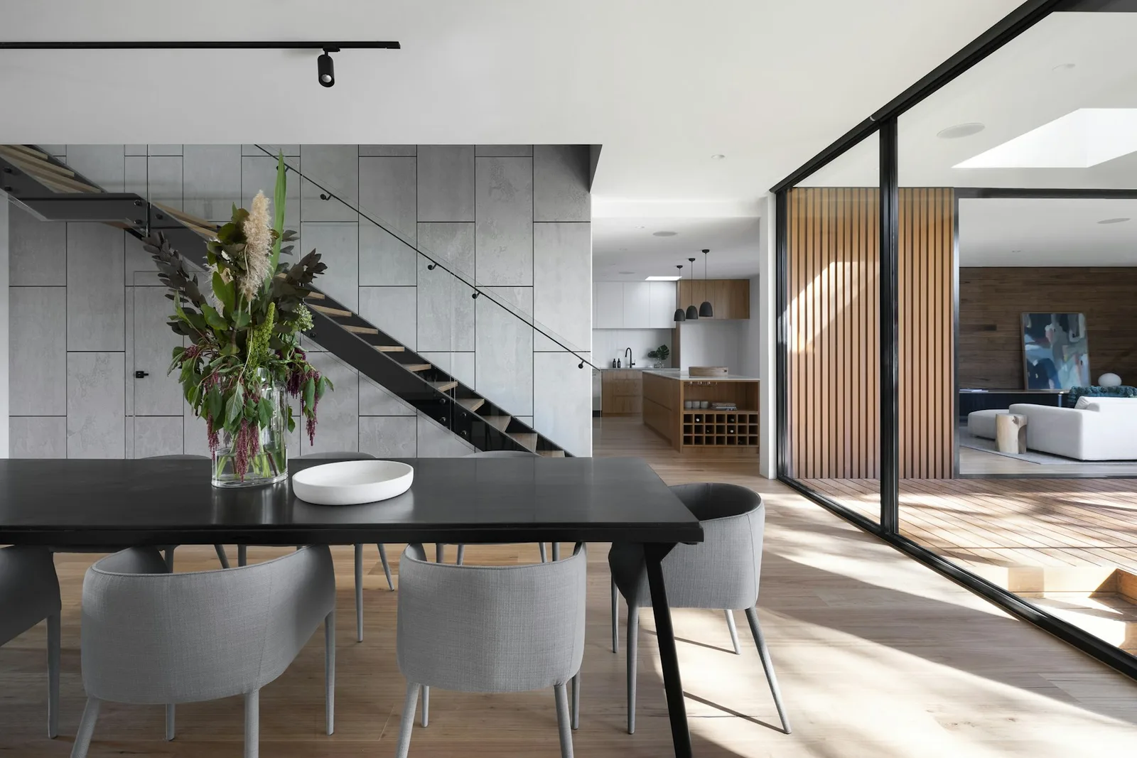 Sunlit dining room and living space with a dark wood table, glass doors, and a sculptural stair